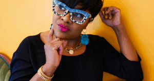 A thoughtful woman wearing bold glasses and jewelry, resting her hand on her chin against a bright yellow background — symbolizing reflection on choosing the right investment.